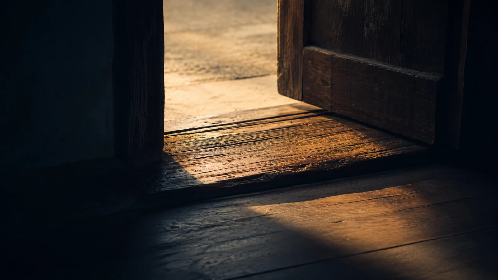 A worn wooden threshold seen from inside a dim room, warm evening light spilling across it from a door left ajar